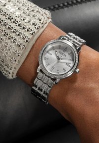 Silberne Guess-Uhr mit rundem Ziffernblatt, Diamantakzenten und einem Mesh-Armband. Das Ziffernblatt weist ein Sonnenstrahlenmuster und rechteckige Stundenmarkierungen auf.