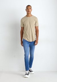 T-shirt en coton beige à manches courtes, assortie à un jean bleu clair et des baskets noir et blanc. Modèle se tenant devant un fond uni.