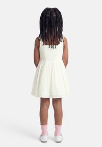 Jeune fille aux cheveux tressés portant une robe blanche sans manches, des chaussettes roses et des chaussures blanches, debout de dos sur un fond uni.