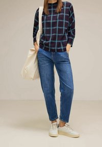 Top a maglia a scacchi blu navy e teal, jeans azzurri chiari e sneakers bianche. Porta una borsa a spalla color crema. Stile casual, vestibilità rilassata.