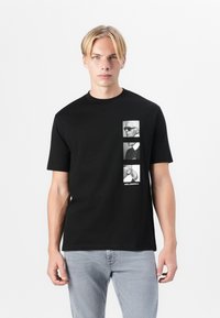 Fekete pamut póló, amelyen három fekete-fehér portré vertikálisan helyezkedik el, alattuk a "KARL LAGERFELD" felirat olvasható.
