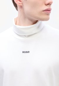 Fehér, puha anyagból készült garbó, fekete "HUGO" logóval a mellkason. Sima textúrájú, magas nyakkal tervezve.