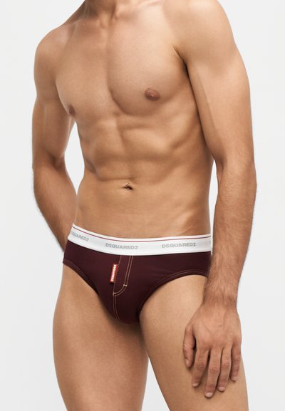 Buste masculin musclé portant un slip bordeaux avec ceinture blanche marqué "DSQUARED2" sur un fond clair uni.