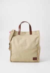 Sac fourre-tout en toile beige avec poignées et bandoulière en cuir marron, comportant une petite pièce en cuir marron avec le logo de la marque. Texture lisse.