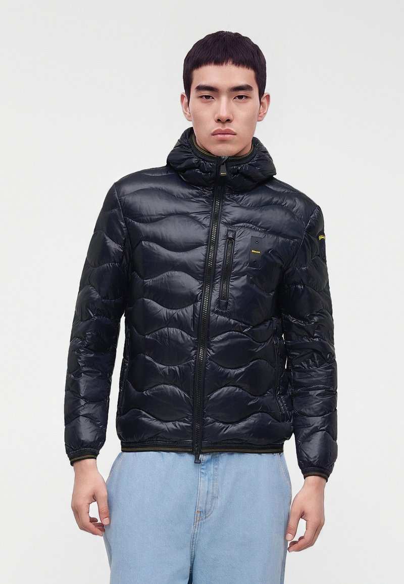 Jeune homme portant une doudoune matelassée noire avec une poche poitrine zippée et un jean bleu clair, debout devant un fond uni.
