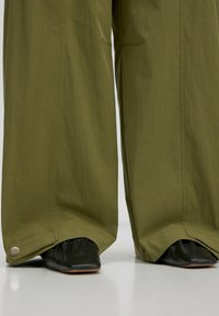 Khaki-weite Hose mit glatter Textur, ausgestattet mit einer Seitentasche und silberner Knopfdetail, kombiniert mit schwarzen flachen Schuhen.