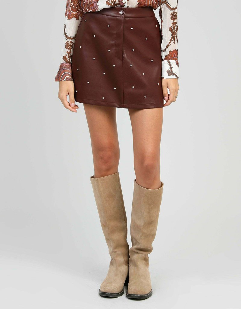 Femme portant une mini-jupe en cuir marron avec des clous en perles, un haut à manches longues à motifs, et des bottes en daim beiges monte-genoux sur un fond gris.