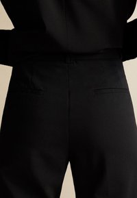 Musta räätälöity housu, jonka pinta on sileä. Housuissa on kaksi sivutaskua, nappilukko ja istuva vyötärö.