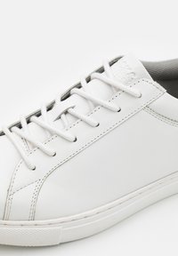 White leather sneaker with flat laces, rounded toe, and contrasting grey inner lining. Features subtle branding and stitched accents.