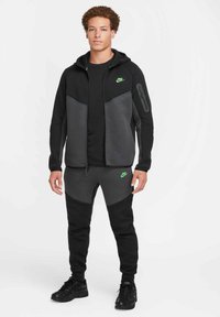 Joven vistiendo un chándal negro y gris oscuro de Nike con logos en verde neón, zapatillas negras, de pie contra un fondo blanco.