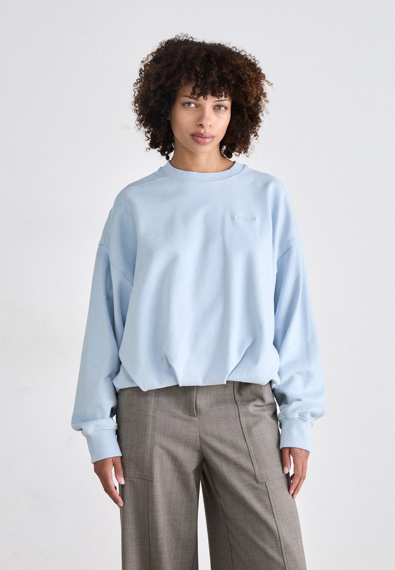 Lyseblå oversized sweatshirt i mykt stoff med rund hals og ribbestrikkede mansjetter, kombinert med grå høyhviste vide bukser.