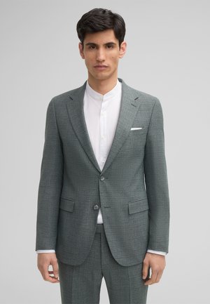 Giacca di abito grigia con texture, colletto a rever, due bottoni e tasche frontali, indossata sopra una camicia bianca con colletto. Vestibilità su misura.