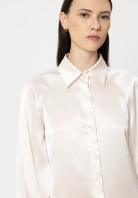 Light ivory, satin blouse with a classic collar, button front, and long sleeves. Features a smooth texture and subtle sheen.