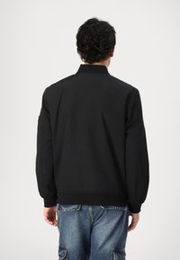 Black bomber jacket with ribbed cuffs and collar. Features a smooth, matte texture. Worn over blue cargo jeans. Minimalistic design.