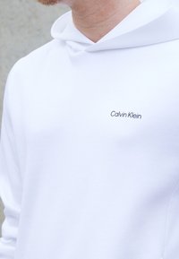 Witte katoenen hoodie met een trekkoordkap en een klein zwart "Calvin Klein" logo op de borst. Gladde textuur en casual ontwerp.