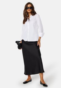 Camicia bianca con bottoni, maxi gonna nera in satin, ballerine nere e una pochette nera con texture. Capelli lunghi, occhiali da sole neri oversize.