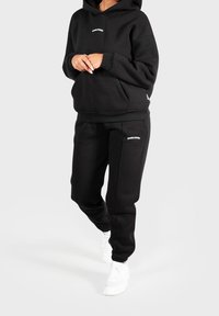 Personne portant un sweat à capuche noir et un pantalon de jogging avec un logo blanc, les mains jointes et des baskets blanches, se tenant devant un fond clair.