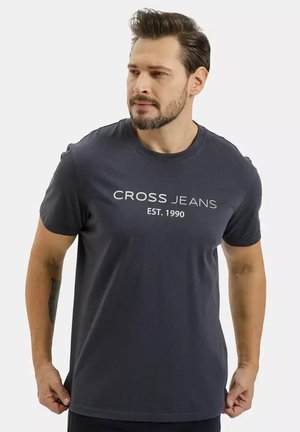 Mężczyzna z brodą, noszący ciemnoszarą koszulkę z napisem "CROSS JEANS EST. 1990", trzyma brzeg koszulki, lekko patrzy na bok.