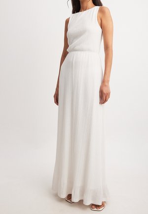 Kobieta w białej, teksturowanej, długiej sukience bez rękawów z dopasowaną talią, stojąca na tle jednolitego, jasnego tła.