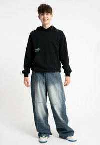 Czarna bluza z kapturem z kieszenią z przodu, z niebieskim logo; zestawiona z jasnoniebieskimi luźnymi dżinsami z bogatym przeszyciem i detalami tekstury.