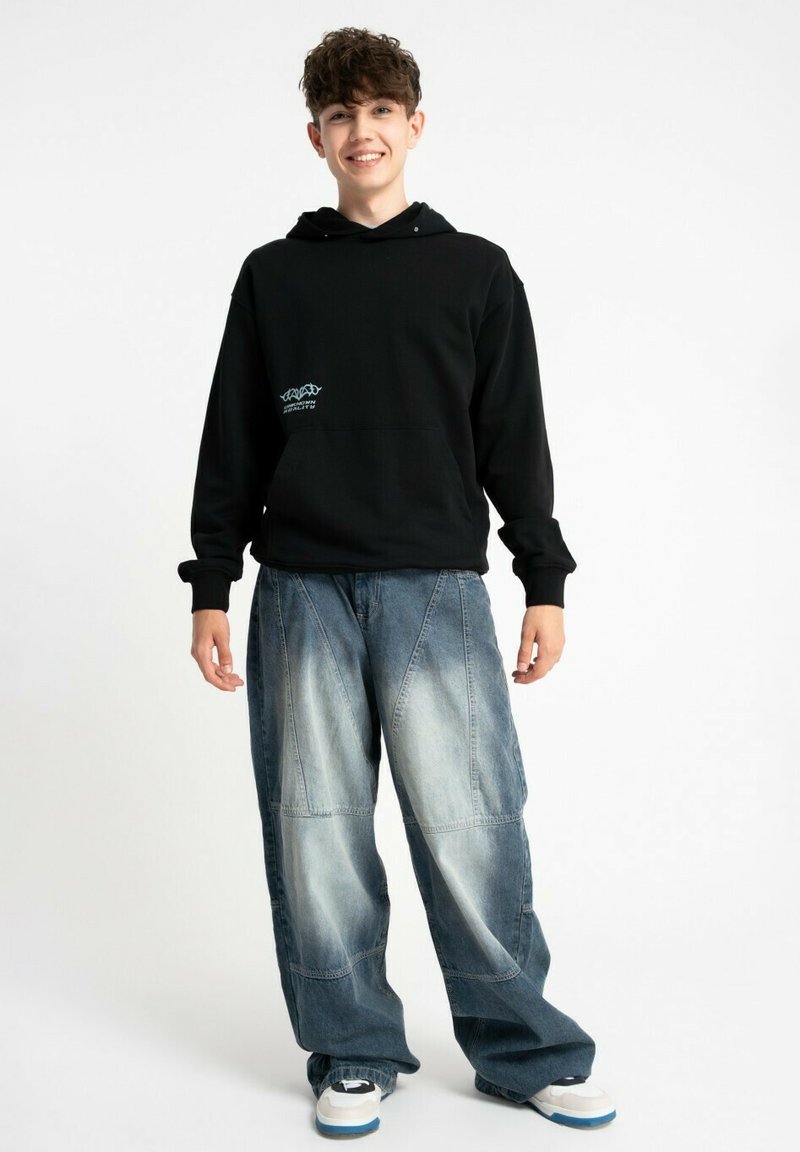 Czarna bluza z kapturem z kieszenią z przodu, z niebieskim logo; zestawiona z jasnoniebieskimi luźnymi dżinsami z bogatym przeszyciem i detalami tekstury.