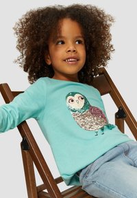 Turquoise lange mouw shirt met een pailletten uil ontwerp in beige, groen en bordeaux. Spijkerbroeken maken de outfit compleet op een houten stoel.