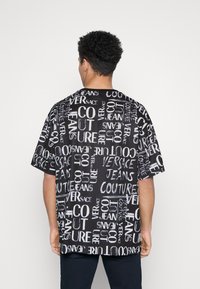 Černé tričko s krátkým rukávem s bílým grafickým vzorem textu "Versace Jeans Couture." Volný střih s kulatým výstřihem.