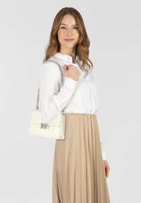 Bolso de cuero sintético blanco con diseño de solapa, accesorios de plata y correa de cadena, combinado con una camisa blanca y una falda plisada beige.