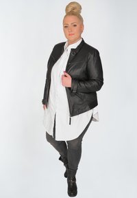 Schwarze Leder-Motorradjacke mit Reißverschluss, kombiniert mit einem weißen Hemd und grauen Hosen, ergänzt durch schwarze Ankle Boots.