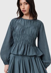 Top peplum in tessuto testurizzato di colore blu ardesia, con dettagli arricciati, maniche lunghe e scollo rotondo. Presenta un corpetto aderente e un orlo svasato.
