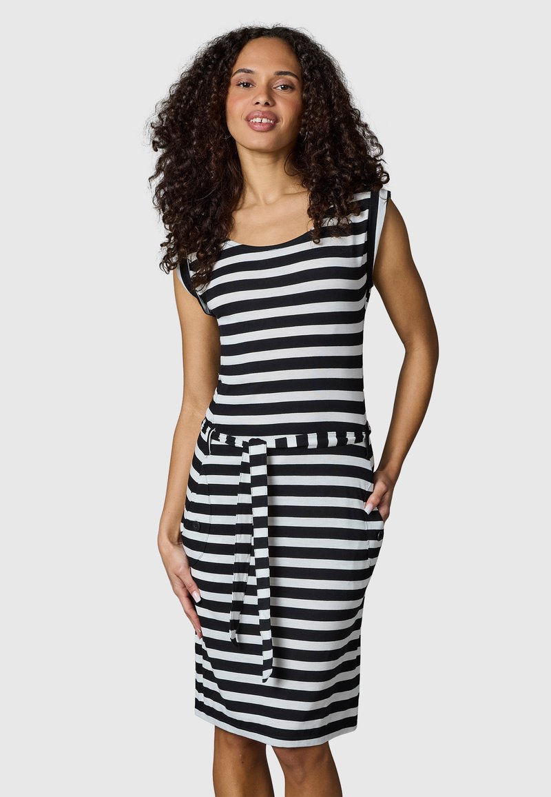 Frau mit lockigem Haar, die ein schwarz-weiß gestreiftes, ärmelloses Kleid mit Taillengürtel trägt, steht vor einem schlichten Hintergrund.
