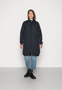 Cappotto lungo imbottito blu navy con colletto alto, chiusura con zip e due tasche laterali, abbinato a jeans blu chiaro e stivali neri.