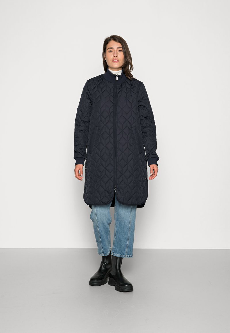 Cappotto lungo imbottito blu navy con colletto alto, chiusura con zip e due tasche laterali, abbinato a jeans blu chiaro e stivali neri.
