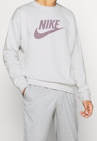 Sudadera gris claro con cuello redondo, puños y cintura acanalados. Presenta un logo de Nike púrpura centrado en el pecho.