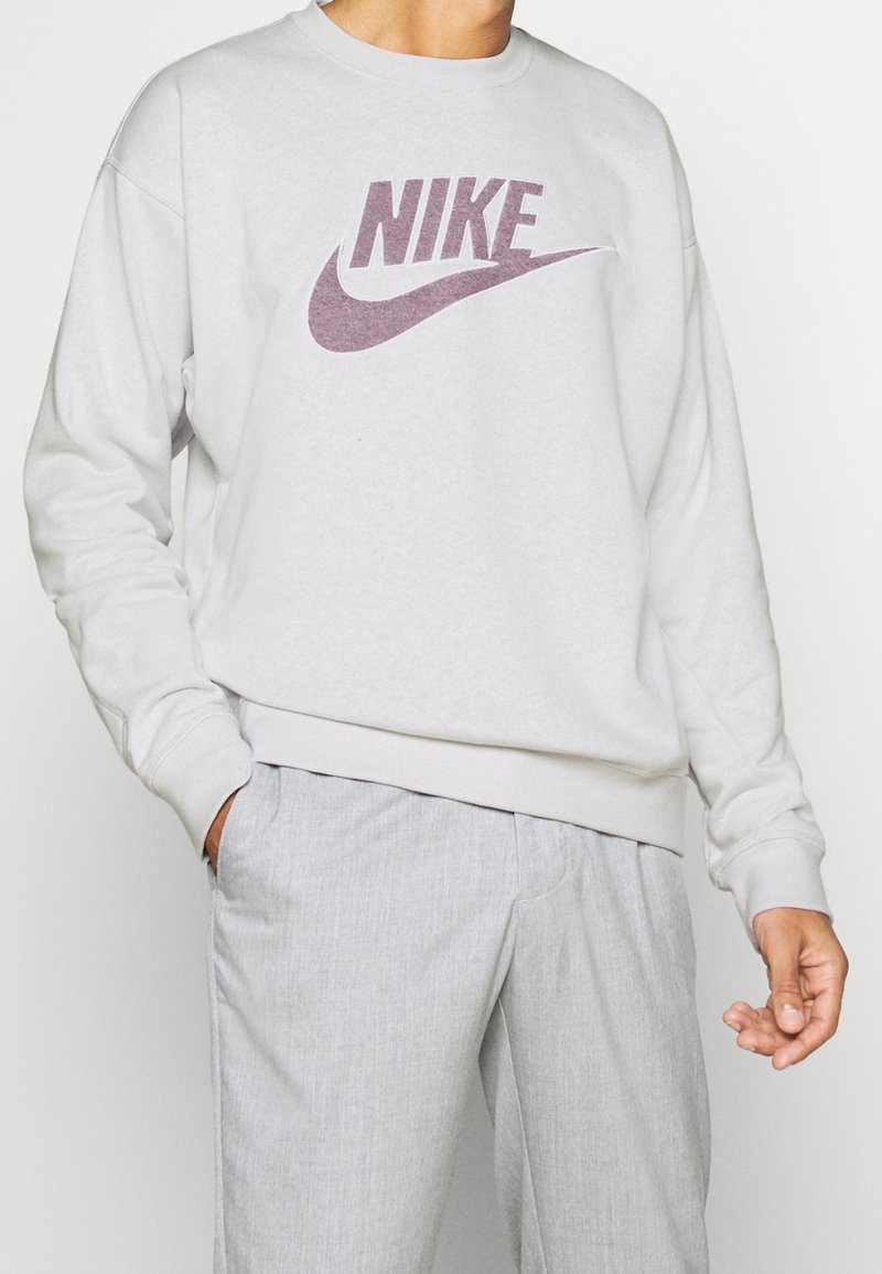 Sudadera gris claro con cuello redondo, puños y cintura acanalados. Presenta un logo de Nike púrpura centrado en el pecho.