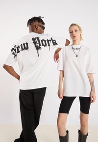 Hvide oversized t-shirts med "New York" i sort dråbe-font på ryggen og forsiden. Sorte bukser og en sølvkæde på modellen foran.