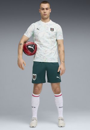 Junger männlicher Fußballspieler steht, hält einen roten Puma-Ball unter dem linken Arm, trägt ein weißes Trikot, dunkelgrüne Shorts und weiße Socken mit Streifen.