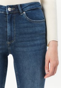 Close-up of a person wearing dark blue denim jeans and a cream ribbed long-sleeve top with hand resting by the side.