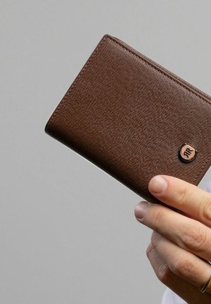 Roka drži zaprto rjavo usnjeno denarnico z teksturirano površino in majhnim kovinskim logotipom na sivi podlagi.