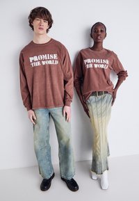 To modeller bærer langermede brune t-skjorter med teksten "PROMISE THE WORLD". Den mannlige modellen kombinerer skjorten sin med lyseblå, vide jeans. Den kvinnelige modellens antrekk har et langt, ombre denimskjørt med en frynset kant.