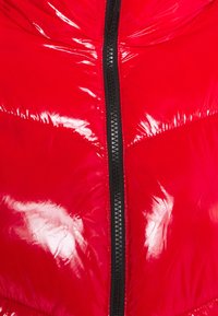 Czerwona puchowa kurtka o błyszczącym wykończeniu, z czarnym zamkiem błyskawicznym i przeszyciami. Materiał wydaje się pikowany, lekki w konstrukcji.