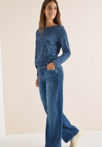 Blau gemusterte Langarmbluse mit Rundhalsausschnitt, kombiniert mit weit geschnittenen blauen Jeans und hellen Sneakers.