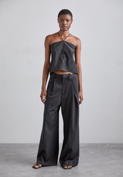 Black cropped top made from smooth material with thin straps; paired with loose black trousers featuring a distinctive front pocket detail.