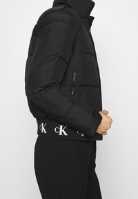 Svart puffjacka med hög krage, sidofickor med dragkedja och elastisk midja med vit branding. Mjuk, quiltad textur synlig.