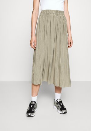 Femme portant une jupe midi plissée beige avec une ceinture élastique, des chaussettes blanches et des baskets de sport noires et grises.
