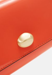 Bolso de mano de charol naranja con cierre redondeado de botón dorado y costuras negras contrastantes a lo largo de los bordes. Textura de superficie brillante.