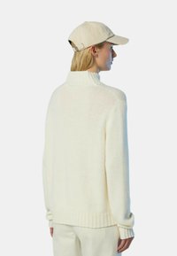 Maglione a maglia color crema con colletto alto e polsini e orlo a coste. Indossato con un berretto beige chiaro, visto da dietro.