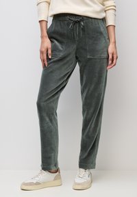 Osoba nosząca zielone spodnie sztruksowe z przednimi kieszeniami i ściągaczem w pasie, w połączeniu z białymi tenisówkami i kremowym swetrem.