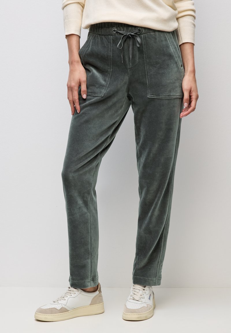 Osoba nosząca zielone spodnie sztruksowe z przednimi kieszeniami i ściągaczem w pasie, w połączeniu z białymi tenisówkami i kremowym swetrem.