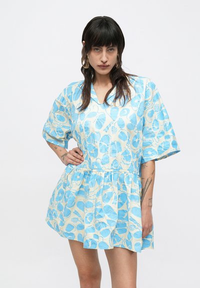 Γυναίκα με σκουρόχρωμα μαλλιά που φοράει ένα κοντό μπλε-λευκό φόρεμα με σχέδιο και μανίκια μέχρι τον αγκώνα, στέκεται με το ένα χέρι στη μέση της μπροστά σε ένα απλό φόντο.